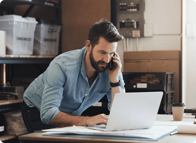 Man bent over talking on a cellphone while tying on his laptop.