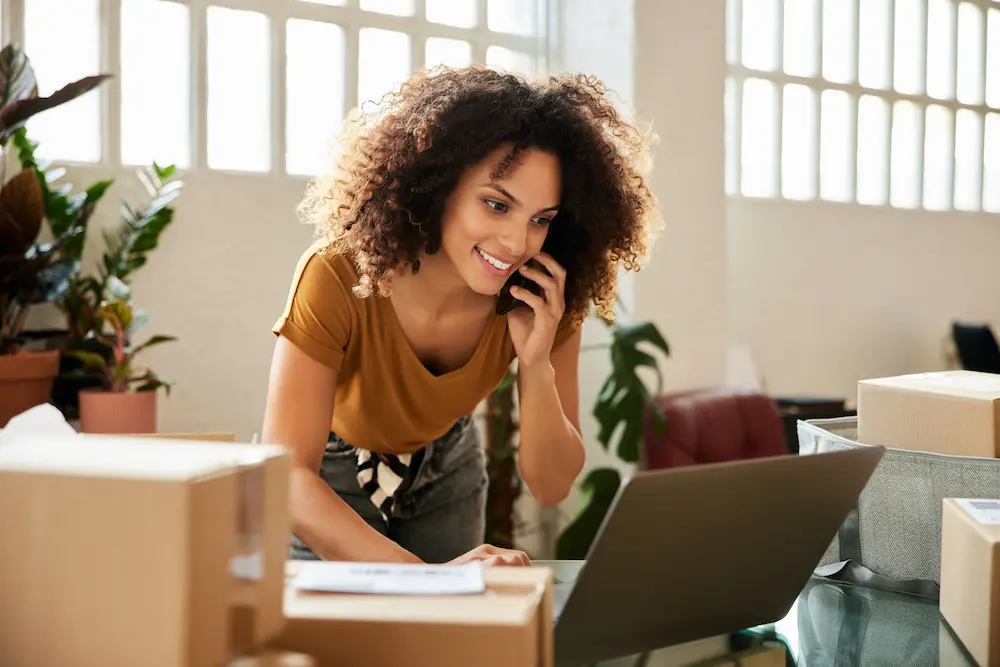 Photo of smiling woman talking on mobile phone while typing on laptop PC.